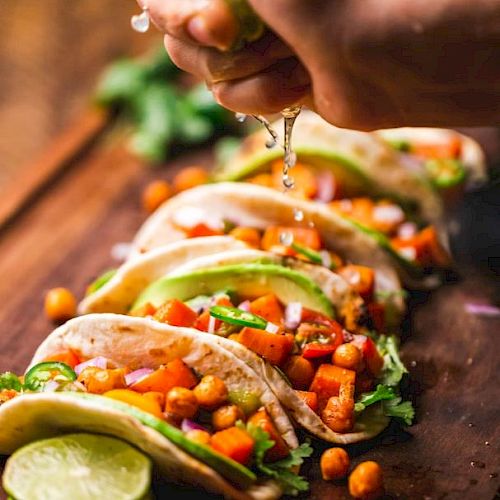 A hand squeezing lime over tacos filled with chickpeas, diced vegetables, and avocado on a wooden surface.