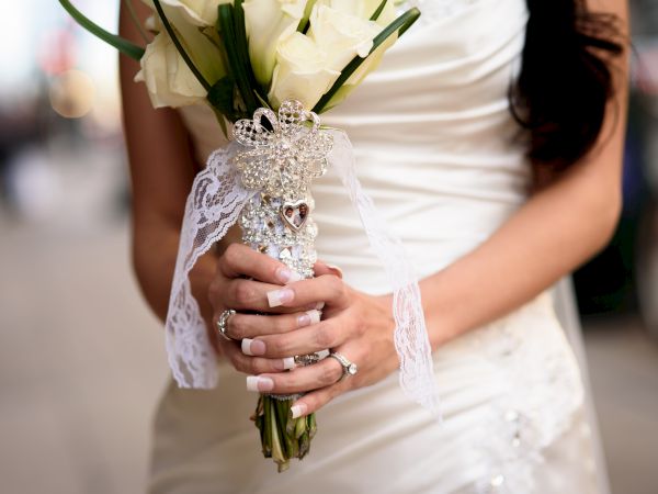 A bride holding a bouquet of white roses, adorned with lace and pearls, wearing a white wedding dress.